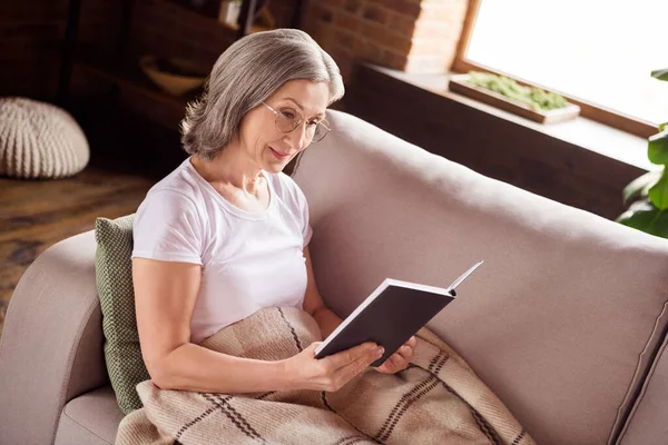 Portrait of attractive focused grey-haired woman sitting on sofa reading interesting book at home house indoors modern brick loft interior — ストック写真