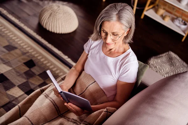 Фото очаровательной пенсионерки носить белую футболку лежа диван улыбаясь читая роман в помещении дома квартиру — стоковое фото