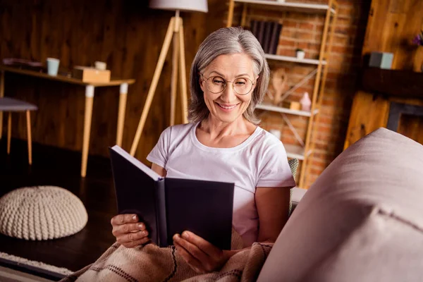 Portrait of attractive cheerful focused grey-haired woman sitting on divan reading interesting book at home house flat indoors — ストック写真