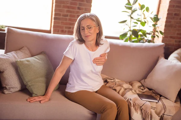 Photo of sad depressed retired woman dressed white t-shirt sitting couch having heart pain arm hand chest indoors flat home house — 图库照片