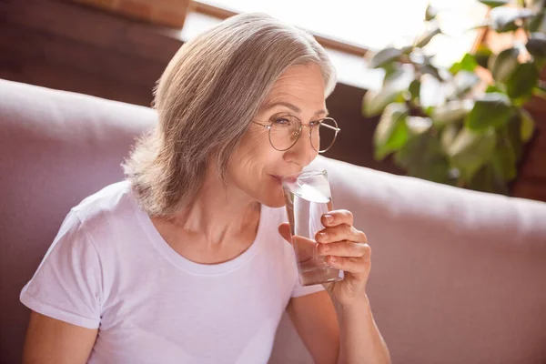 Photo of happy adorable retired woman dressed white t-shirt sitting couch drinking water smiling indoors flat home house — ストック写真