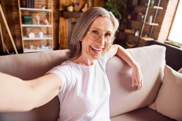 Foto de encantador doce aposentado mulher vestida branco t-shirt sentado sofá gravação vídeo sorrindo dentro de casa plana casa — Fotografia de Stock