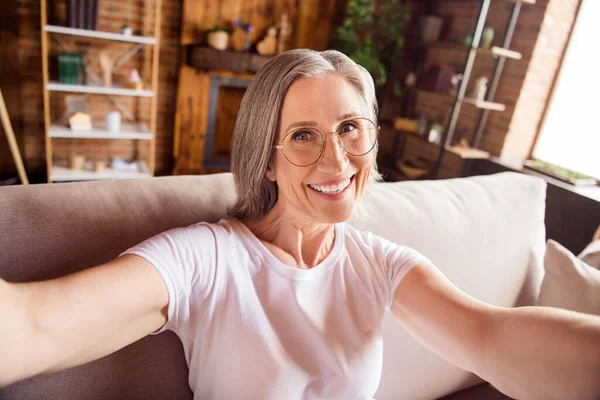 Self-portrait of attractive cheerful grey-haired woman sitting on sofa free spare time at home house indoors modern brick interior — ストック写真