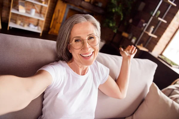 Self-portrait of attractive cheerful grey-haired woman sitting on divan welcoming you at loft industrial home house flat indoors —  Fotos de Stock