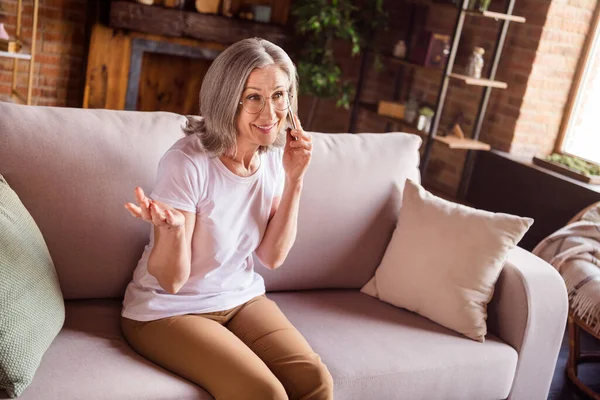 Фото солодкої блискучої жінки на пенсії, одягненої в білу футболку, яка сидить на дивані, розмовляє сучасним пристроєм, посміхаючись у приміщенні житлового будинку — стокове фото