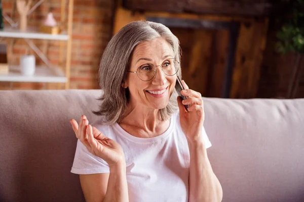 Fénykép aranyos bájos hölgy nyugdíjas viselni fehér póló ül kanapé mosolygós beszél modern szerkentyű beltéri lakás — Stock Fotó