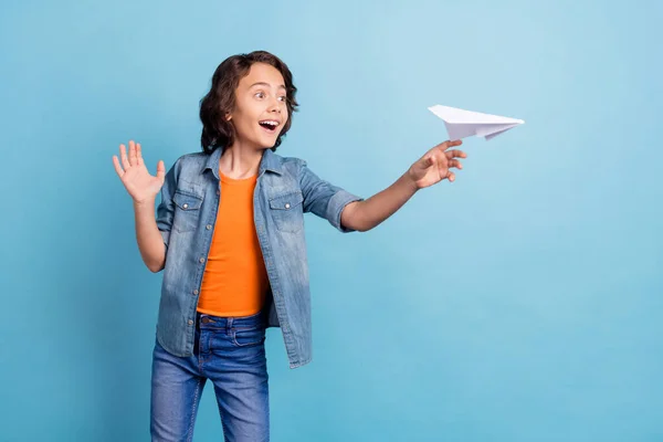 Fotografie mladého vzrušený školní chlapec šťastný pozitivní úsměv hodit létat vzduch papír letadlo izolované přes modré barvy pozadí — Stock fotografie