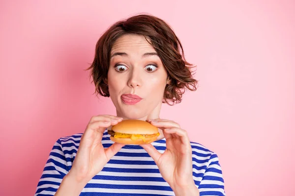 Portré fiatal vonzó funky éhes gyönyörű lány tartsa hamburger nyalás ajkak elszigetelt rózsaszín szín háttér — Stock Fotó