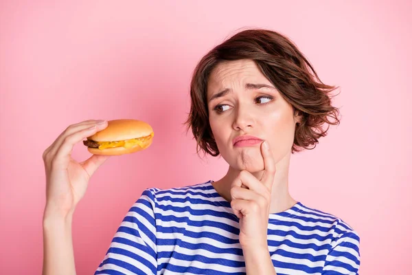 Retrato de joven hermosa chica seria pensativa mirada en hamburguesa con queso mantenga el mentón del dedo aislado en el fondo de color rosa — Foto de Stock