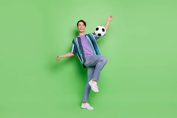 Full size photo of cheerful active joyful happy man kick football smile game isolated on green color background — стоковое фото