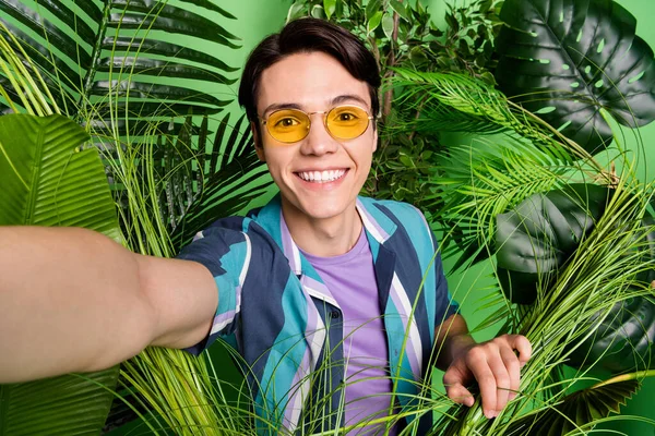 Fotografie veselý mladý šťastný muž nosit sluneční brýle, aby selfie v křoví listy izolované na zeleném pozadí — Stock fotografie