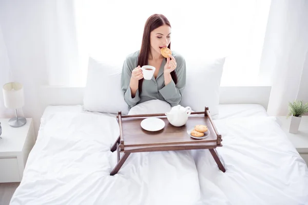 Fotografie docela roztomilé mladé dámy šedé spacáky ležící postel pití ranní káva jíst sušenky s úsměvem v interiéru pokoj domácí dům — Stock fotografie