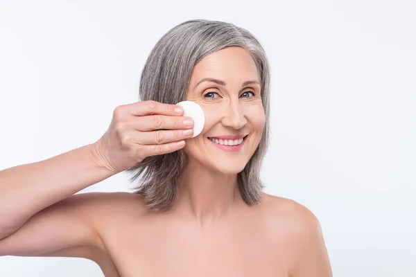 Foto de atraente velha senhora positiva segurar almofada de algodão remover maquiagem sorriso isolado no fundo de cor cinza — Fotografia de Stock