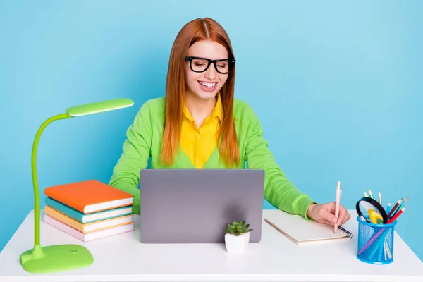 Фото смешной рыжей прической юная студентка писать блокнот смотреть ноутбук носить очки кардиган работы дома изолированы на синем фоне — стоковое фото