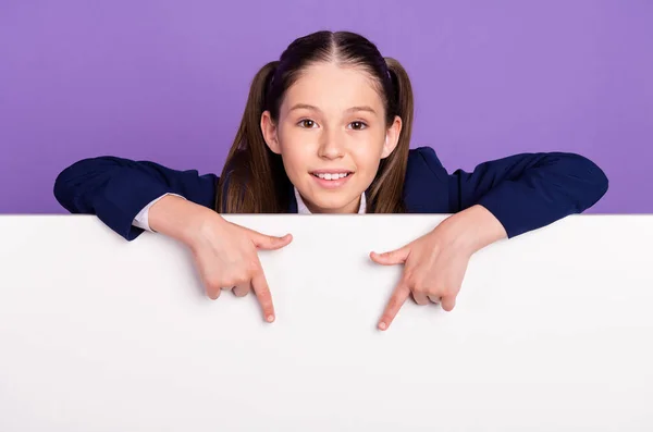 Фото милая очаровательная школьница одета в синюю униформу улыбаясь указывая пальцами белый плакат пустое пространство изолированный фиолетовый цвет фона — стоковое фото