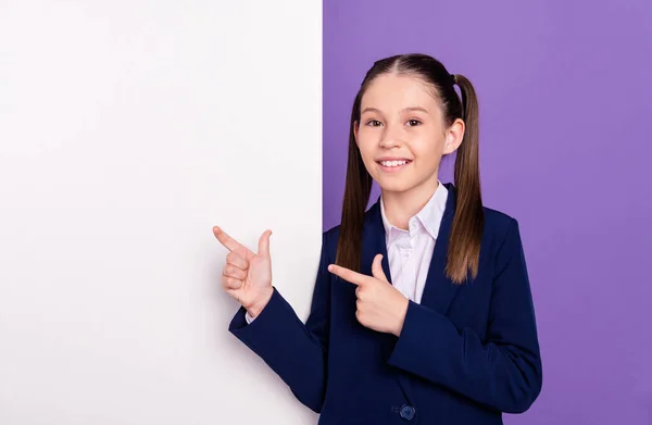 Kuva melko ihana koulutyttö pukeutunut sininen univormu osoittaa sormella valkoinen kyltti tyhjä tila eristetty violetti väri tausta — kuvapankkivalokuva