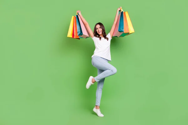 Full size photo of nice brunette young lady hold bags wear t-shirt jeans isolated on green color background — Stock Photo, Image