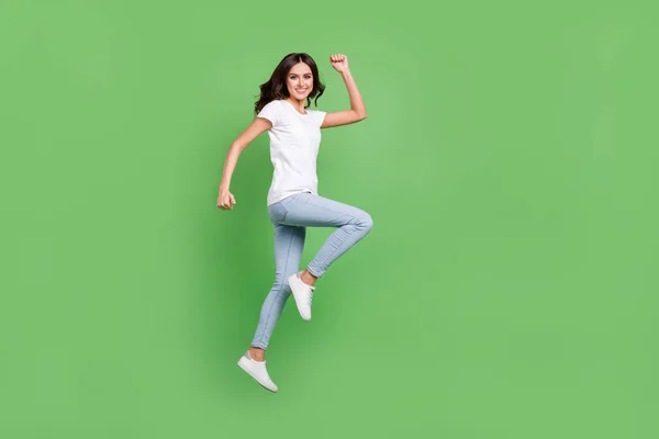 Full size profile photo of cool brunette young lady jump hands fists wear t-shirt jeans isolated on green color background — Stock Photo, Image
