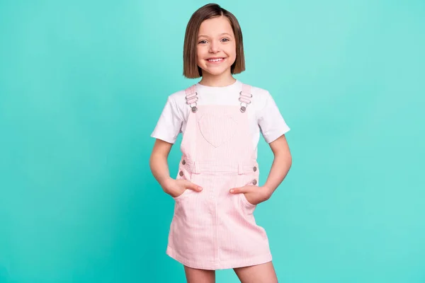 Photo of cheerful happy small positive girl hold hands pockets smile pink dress isolated on teal color background