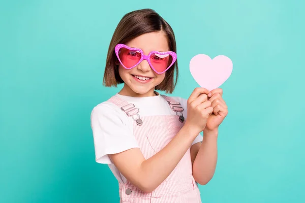 Foto de engraçado adorável menina da escola usar rosa óculos gerais sorrindo segurando pouco coração isolado fundo cor teal — Fotografia de Stock