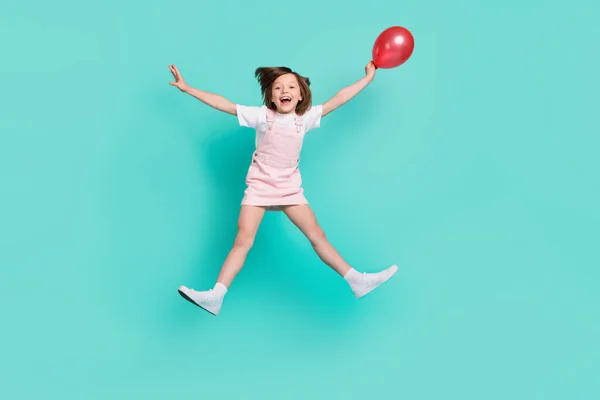 Foto van grappig schattig schoolmeisje gekleed roze overall springen hoog houden rode ballon glimlachen geïsoleerde turquoise kleur achtergrond — Stockfoto