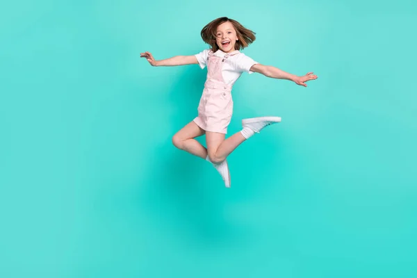 Foto de adorable brillante niña de la escuela usar rosa en general sonriendo saltando los lados de los brazos altos fondo de color verde azulado aislado —  Fotos de Stock