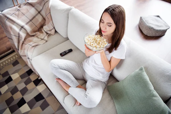 Foto retrato morena mulher sentada no sofá em casa assistindo tv cheirando milho pop — Fotografia de Stock