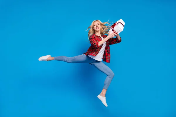 Photo of impressed cute young woman dressed checkered shirt jumping high holding gift box smiling isolated blue color background