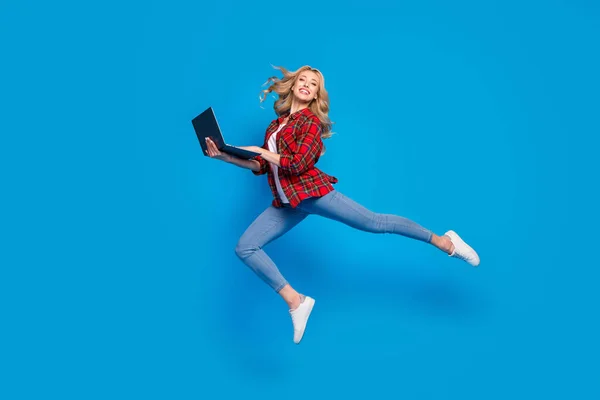 迷人甜美的年轻女士穿着格子花衬衫微笑跳跃着拿着现代小玩意孤立的蓝色背景的照片 — 图库照片