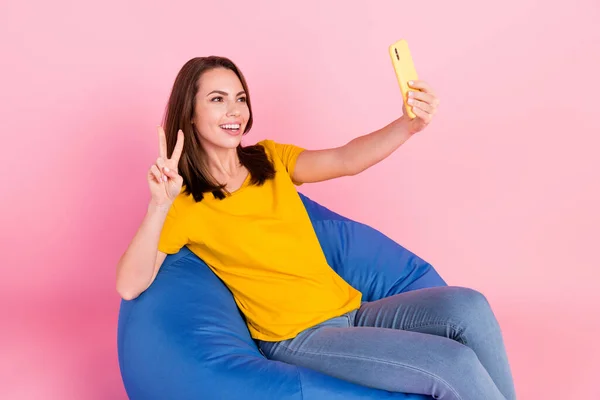 Foto de la fresca morena cabello millennial lady sit do selfie show v-sign wear yellow t-shirt jeans isolated on pink background — Foto de Stock