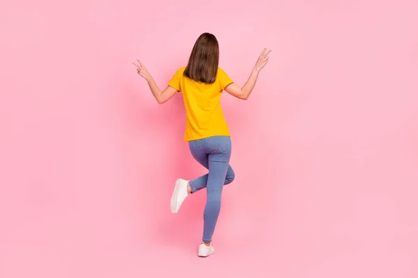 Foto de espalda larga de cabello moreno funnky milenial lady stand show v-sign wear yellow t-shirt jeans isolated on pink color background —  Fotos de Stock