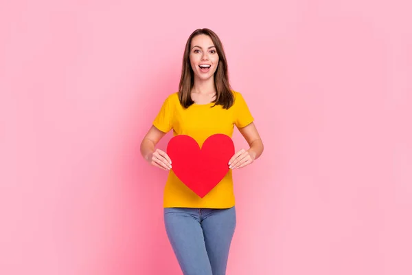 Photo of excited funky young lady wear yellow t-shirt smiling holding red heart isolated pink color background — Stock Photo, Image