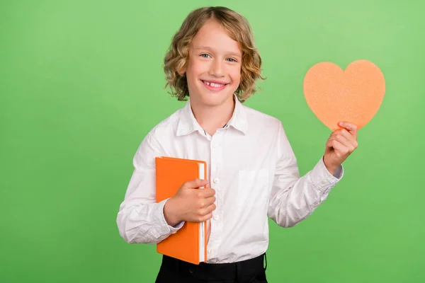 Φωτογραφία του μικρού funky ξανθό αγόρι κρατήσει το βιβλίο καρδιά φορούν λευκό πουκάμισο απομονώνονται σε πράσινο φόντο χρώμα — Φωτογραφία Αρχείου