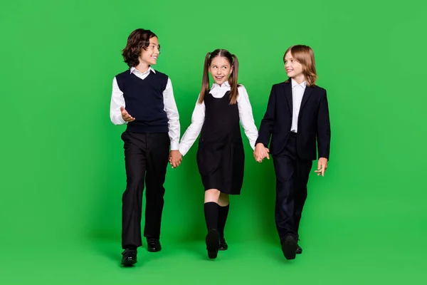 Foto de cuerpo completo de chicos divertidos niñas pequeñas cogidas de la mano van a usar uniforme escolar aislado sobre fondo verde — Foto de Stock