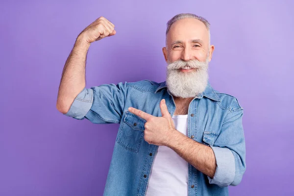 건강 한 노인의 사진 청바지를 입고 손가락으로 근육을 가리키며 보라색 배경을 웃는 모습 — 스톡 사진