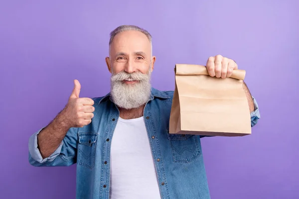 Photo of lucky charming mature man dressed denim outfit smiling showing thumb up holding delivery isolated purple color background