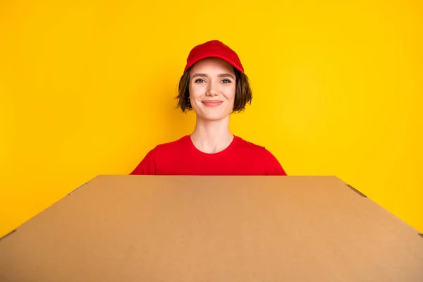 Фотографічний портрет жінки-працівниці в червоній уніформі, що тримає картонну коробку посміхаючись ізольовано на яскраво-жовтому тлі — стокове фото