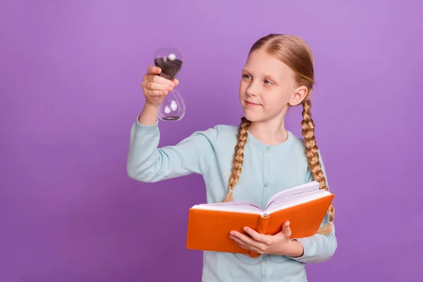 Photo of cute nice pupil girl hold hourglass look read book wear blue shirt isolated violet color background — Stock Photo, Image