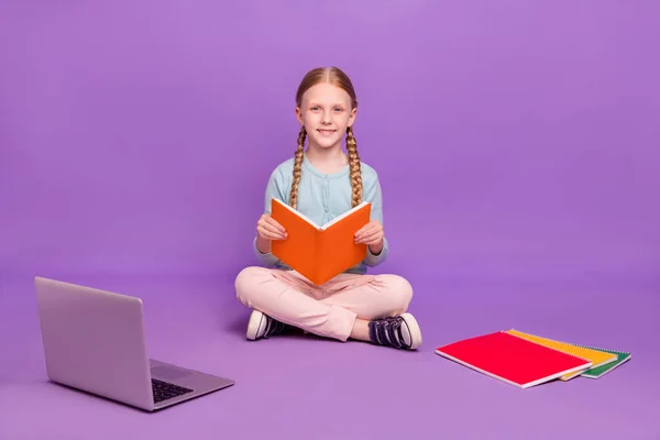 Photo of cheerful positive learner girl sit floor read book toothy smile wear blue shirt isolated purple color background — Stock Fotó