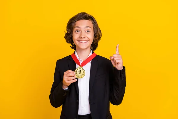 Foto des Genies Gewinner Junge zeigt den ersten Platz Medaille heben Finger tragen schwarze Uniform isoliert gelbe Farbe Hintergrund — Stockfoto