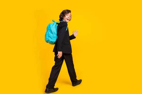 Photo of friendly little schoolboy walk wave palm wear backpack black uniform shoes isolated yellow color background — Fotografia de Stock
