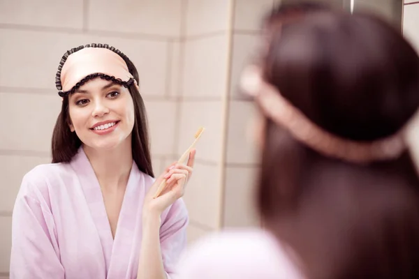 Foto af yndig skinnende ung kvinde lyserød natbeklædning ser spejl børste tænder smilende indendørs hjem værelse - Stock-foto