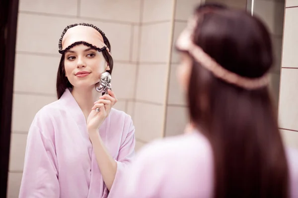 Foto af charmerende smuk ung dame lyserød sleepwear ser spejl smilende massage ansigt indendørs værelse hjem - Stock-foto