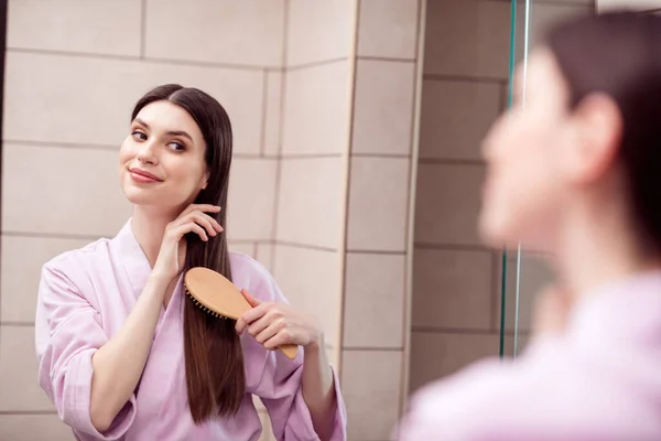 Foto af charmerende sød ung dame lyserød badekåbe sleepwear leder spejl børstning hår indendørs værelse hjem hus - Stock-foto