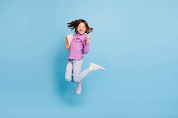 Foto von glänzenden Glück Schulmädchen tragen lila Rollkragen springen halten Werbebücher steigende Faust lächelt isoliert blaue Farbe Hintergrund — Stockfoto