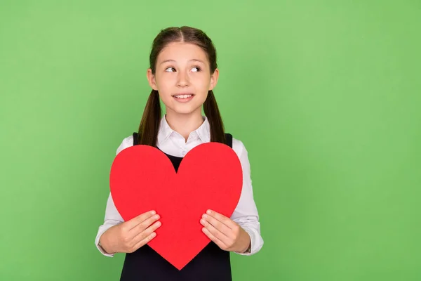 Foto de menina da escola feliz sorriso positivo segurar grande vermelho papel coração sonho olhar vazio espaço isolado sobre cor verde fundo — Fotografia de Stock