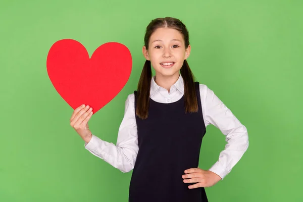 Foto de menina da escola feliz sorriso positivo segurar coração figura sentimentos cartão isolado sobre cor verde fundo — Fotografia de Stock