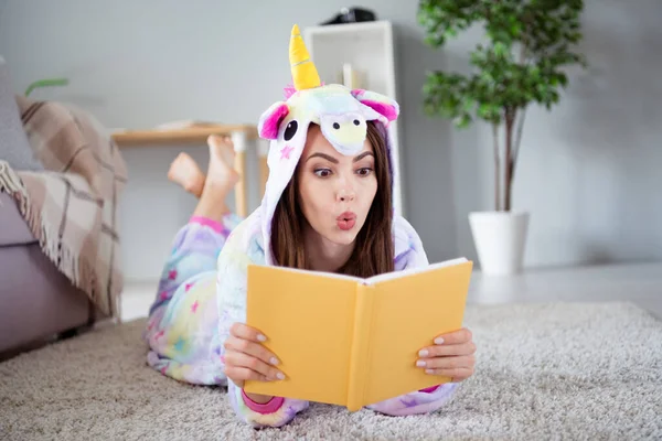 Foto de sorprendido emocionado sorprendido señora infantil laico piso leer libro interesante desgaste acogedor unicornio kigurumi en el interior — Foto de Stock