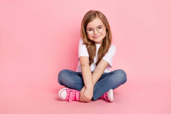 Zdjęcie cute lovely little girl siedzieć podłogi ręce trzymać nosić specyfikacje biały t-shirt dżinsy buty odizolowany różowy kolor tło — Zdjęcie stockowe