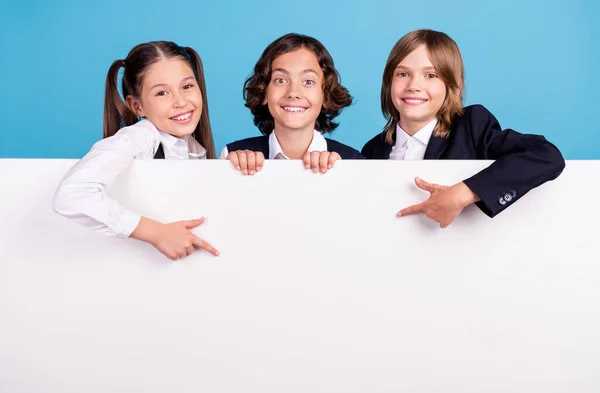 Фото смішних чарівних школярів в уніформі, що тримає вказівний палець білий щит порожній простір ізольований синій колір фону — стокове фото